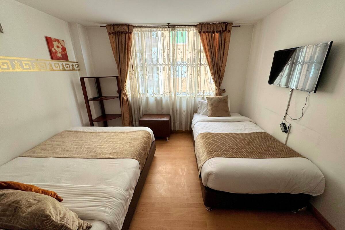 A hotel room with two single beds, a small wooden bench, a television mounted on the wall, and a window with curtains.