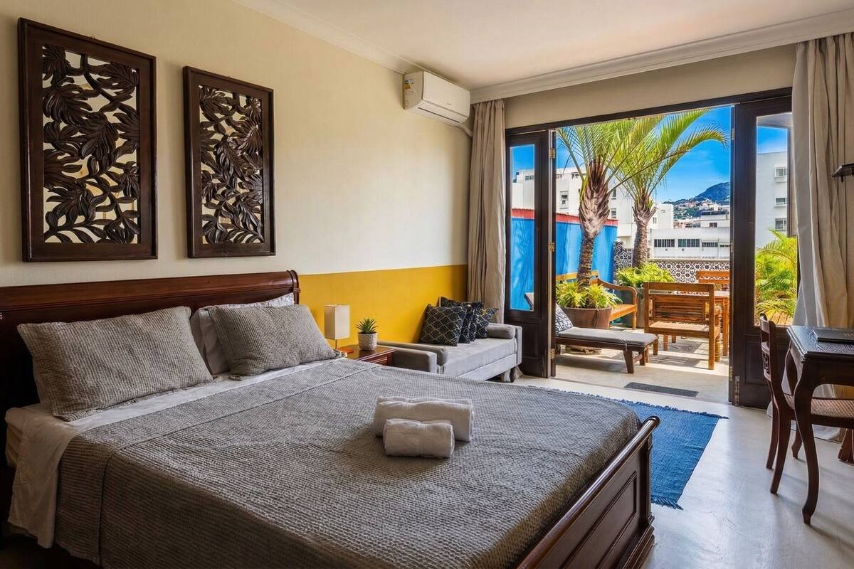 Habitación de hotel con cama, escritorio, silla y balcón con plantas y vistas a edificios.