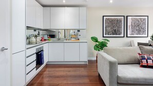 A modern kitchen with white cabinets, a built-in oven, and a countertop with various appliances and a kettle. There is a grey sofa with a checkered pillow and a potted plant in the living area. Two abstract paintings are hung on the wall. - Chancery Lane Apartments (London)
