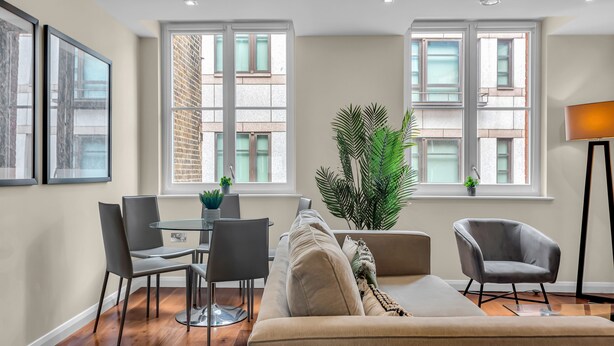 A modern living room with a sofa, chairs, a dining table, and large windows offering a view of a brick building. - Chancery Lane Apartments (London)