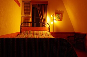 Une pièce faiblement éclairée, avec un lit, une fenêtre avec des rideaux, une chaise et un luminaire fixé au mur.