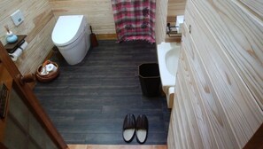 Une petite salle de bain comprenant des toilettes, un bidet, une poubelle et une paire de chaussons posés sur le sol.