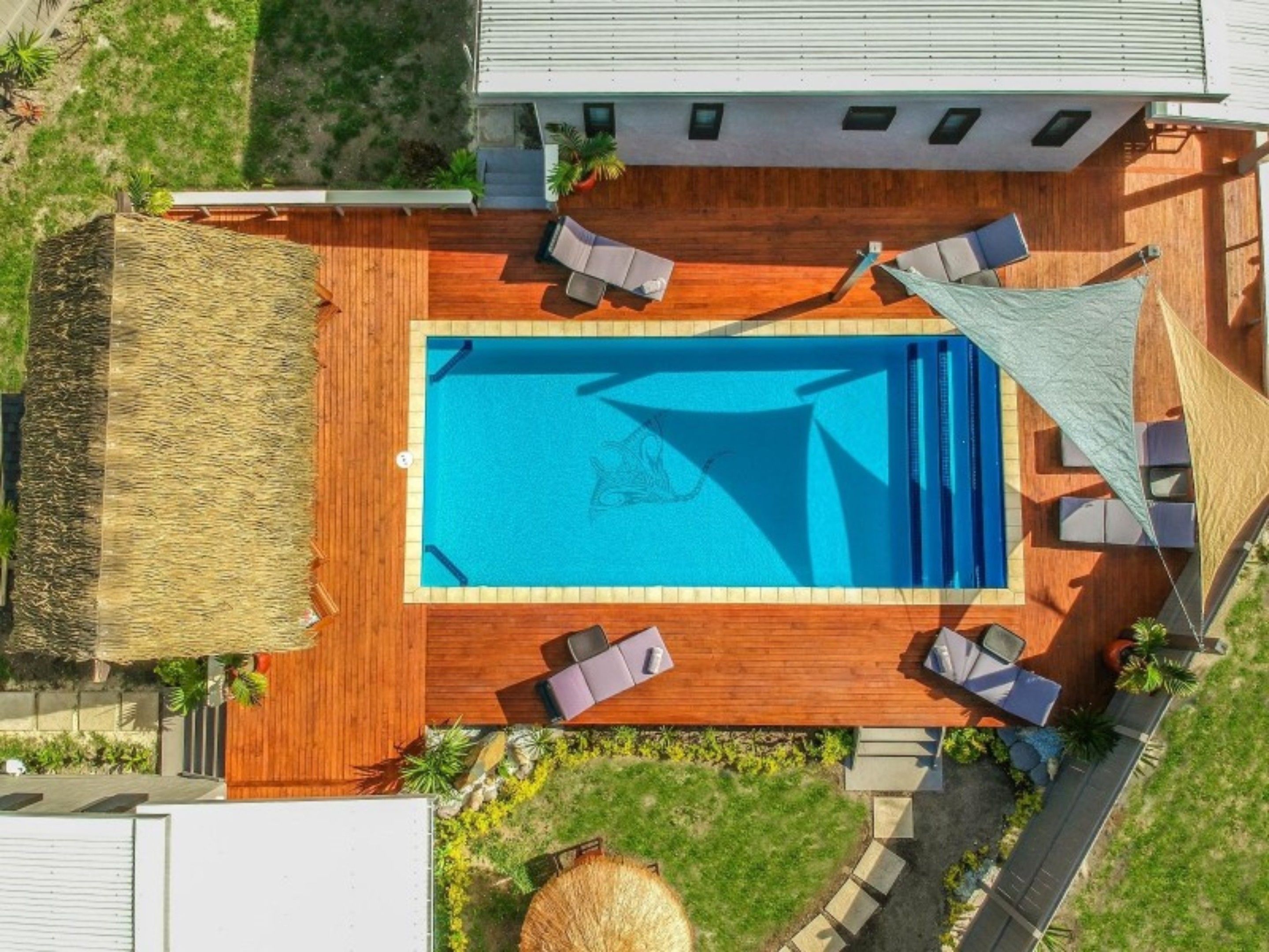 Eine Vogelperspektive auf einen Swimmingpool mit einer Holzterrasse, Liegestühlen und einer Struktur mit Reetdach.