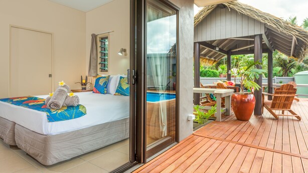 A hotel room with a bed, a sliding glass door leading to an outdoor deck with a pool, and a thatched-roof structure. - Cook Islands Holiday Villas Turangi (Rarotonga)