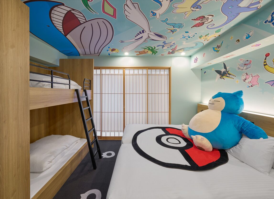 Quarto com beliche, um brinquedo de pelúcia grande, parede com vários adesivos e janela com persianas.