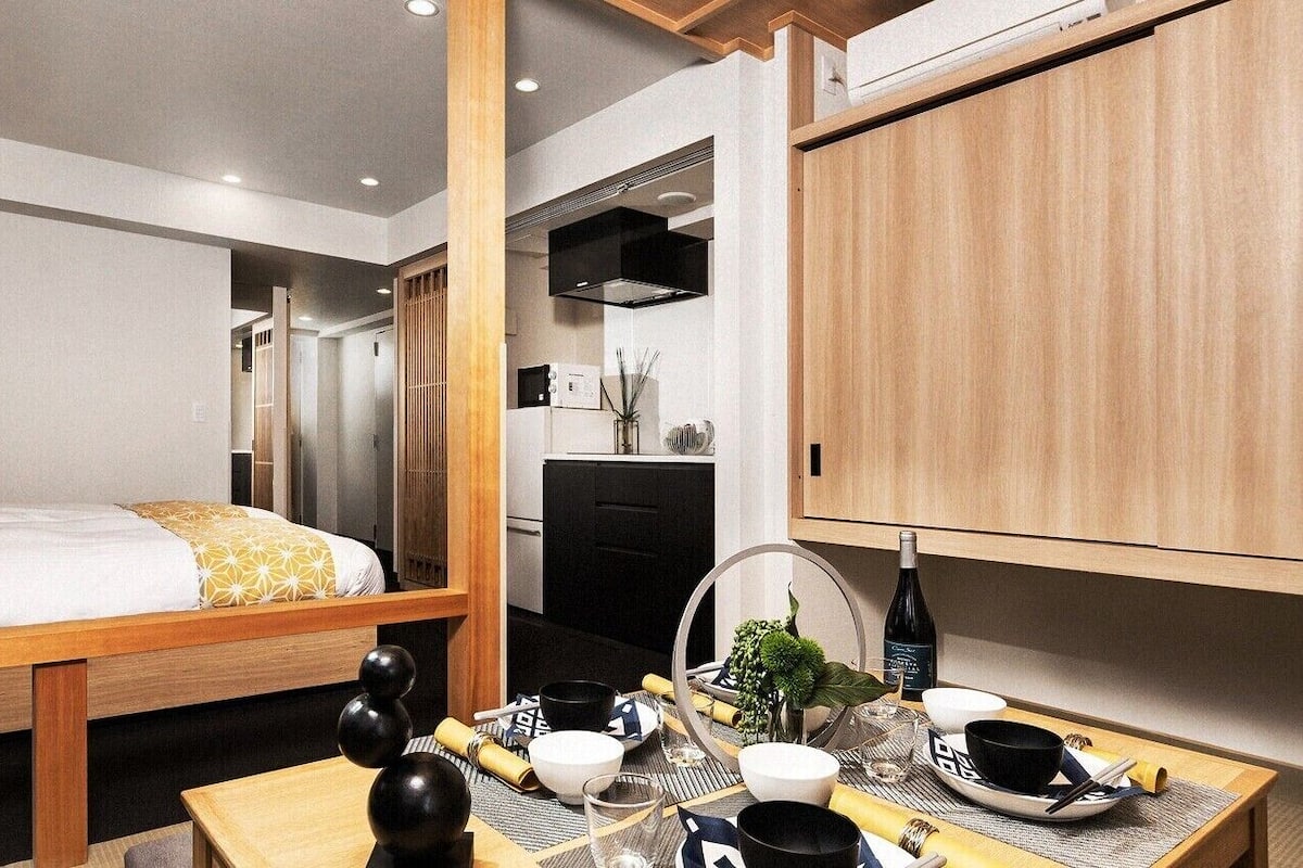 Kompakti hotellihuone, jossa on sänky, pieni ruokapöytä, pieni keittiö ja puinen kaappi.