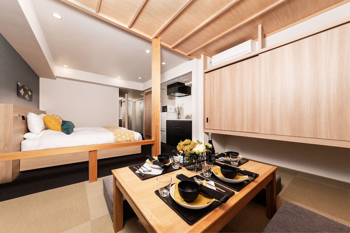 A modern hotel room with a bed, a dining area with a table set for two, and a kitchenette in the background.