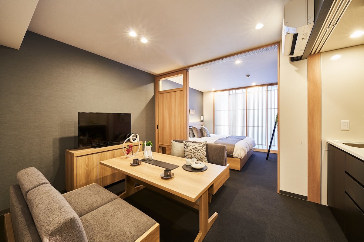 A compact hotel room with a wooden dining table, a sofa, a TV, a bed, and a large window.