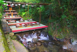 Uma área de refeições ao ar livre tranquila, com mesas e bancos de madeira, instalada ao longo de um riacho com cachoeira.