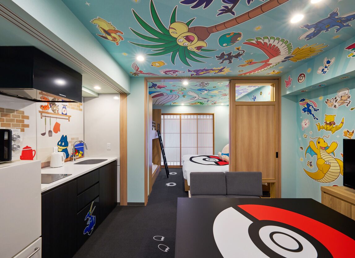 Une chambre d’hôtel dotée d’une kitchenette, d’un lit recouvert d’une grande couverture aux motifs de Pokémon, et d’une fresque murale au plafond représentant différents personnages de Pokémon.