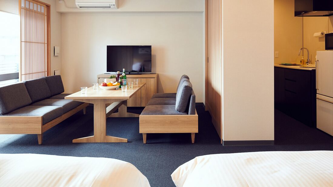 A hotel room with a sofa, a dining table with a fruit bowl, a television, and a kitchenette.
