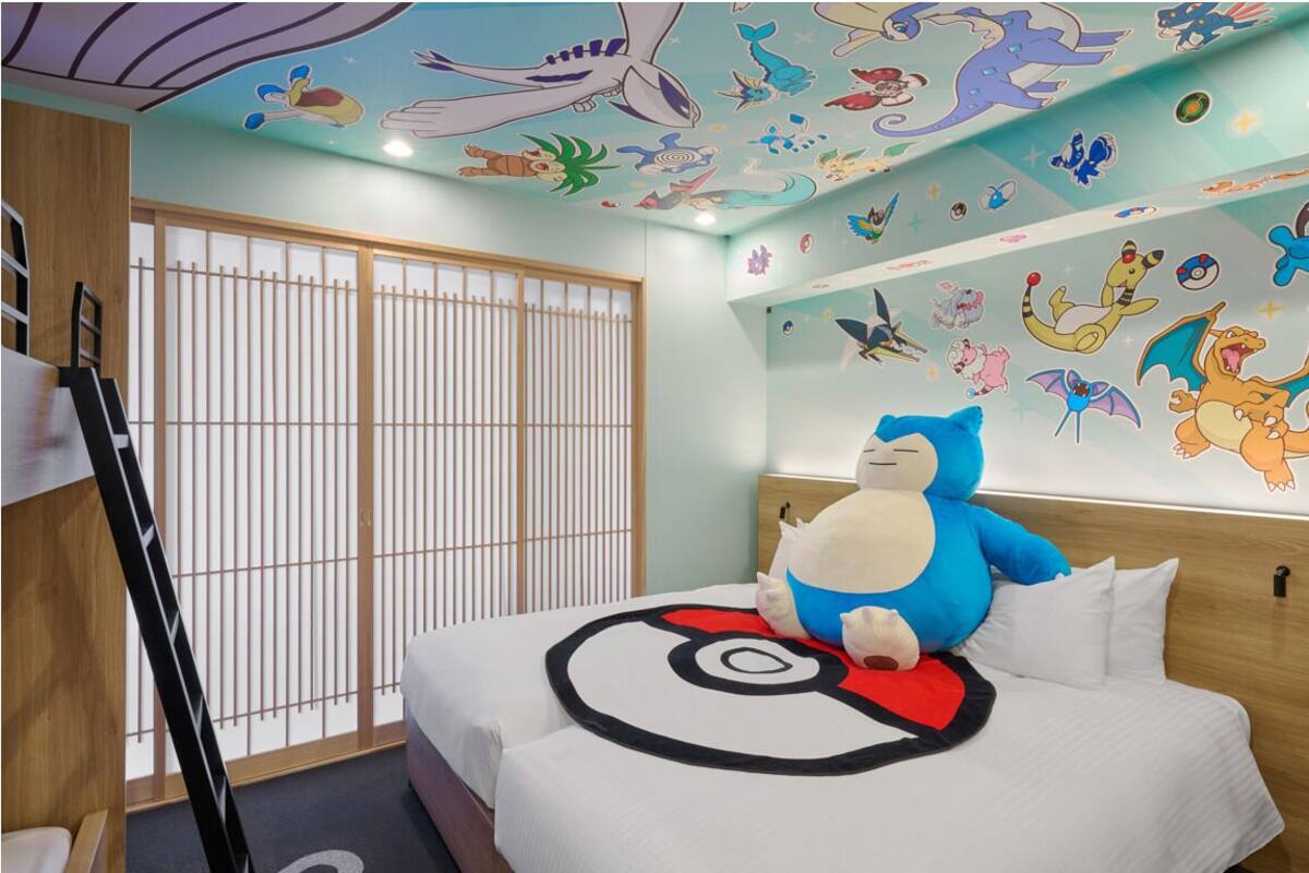 A hotel room with a bed featuring a Pokémon-themed bedspread, a large plush toy, and a wall decorated with various Pokémon characters.