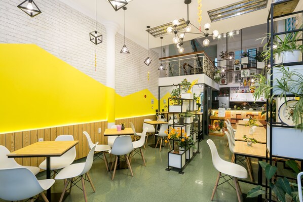 Un café moderno con mesas de madera y sillas blancas, una pared de acento amarilla y diversas plantas.