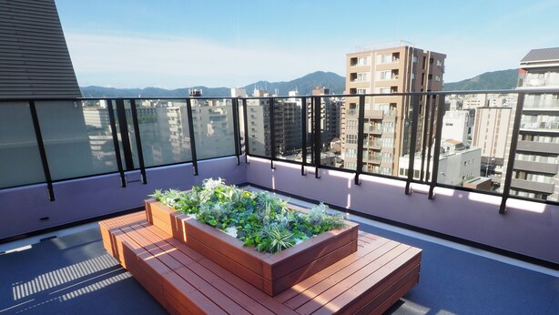 A rooftop terrace with a wooden planter box filled with plants, a seating area, and a view of surrounding buildings and mountains.