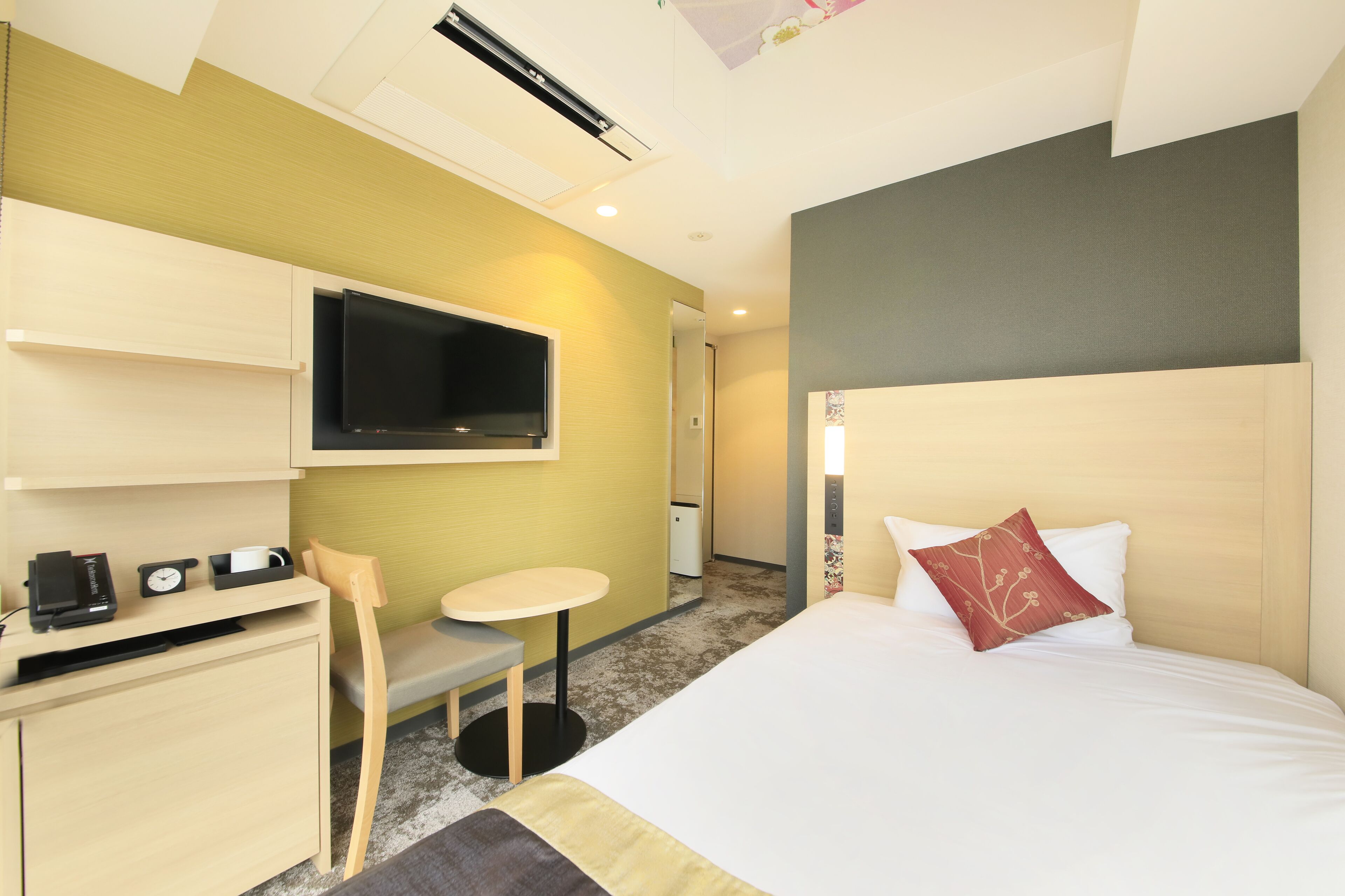 A hotel room with a bed, a desk, a chair, a small table, a TV, and a bathroom visible in the background.