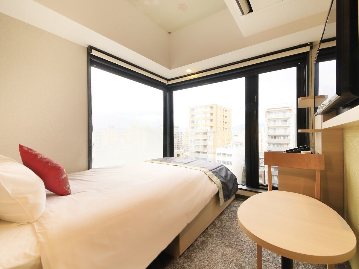 A hotel room with a bed, a small round table, a wardrobe, and large windows offering a city view.