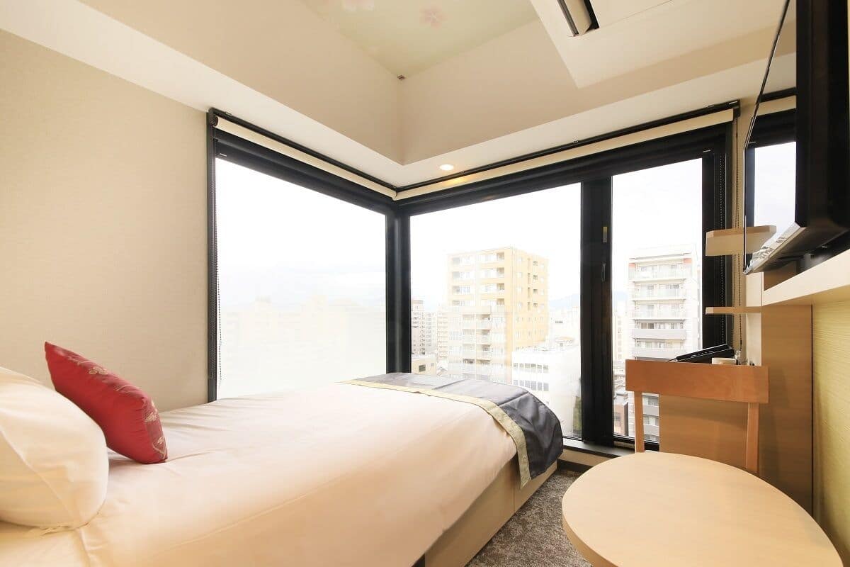 A hotel room with a bed, a small round table, a wardrobe, and large windows offering a city view.