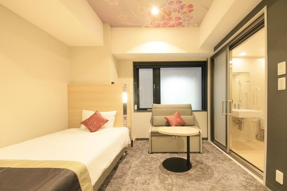A hotel room with a bed, a sofa, a small table, and a glass shower area.