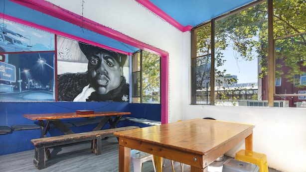 A room with a wooden table, benches, and stools. There is a large mural on the wall and a window with a view of trees. - Hump Backpackers (Potts Point)