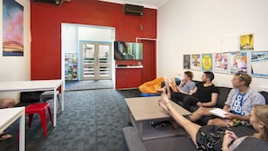 A room with a red wall, a television mounted on the wall, and a group of people sitting on a couch. - Hump Backpackers (Potts Point)