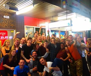 A group of people in a brightly lit indoor space with a metal ceiling and a sign indicating a 7-day open 24 hours service. - Hump Backpackers (Potts Point)