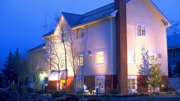 A two-story house with a brick chimney, a porch with chairs, and a small tree with lights. - Melange (Fujikawaguchiko)