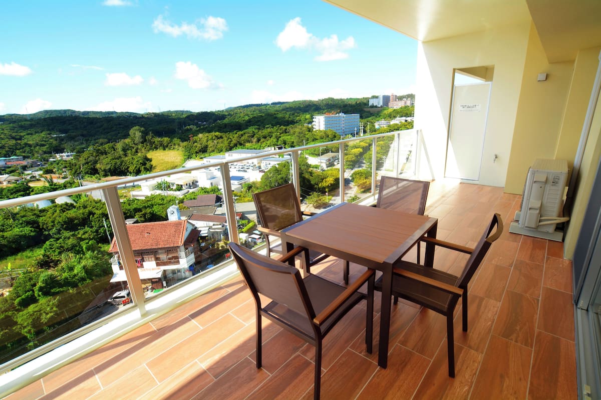 A balcony with a wooden table and chairs, offering a view of a green landscape and buildings.
