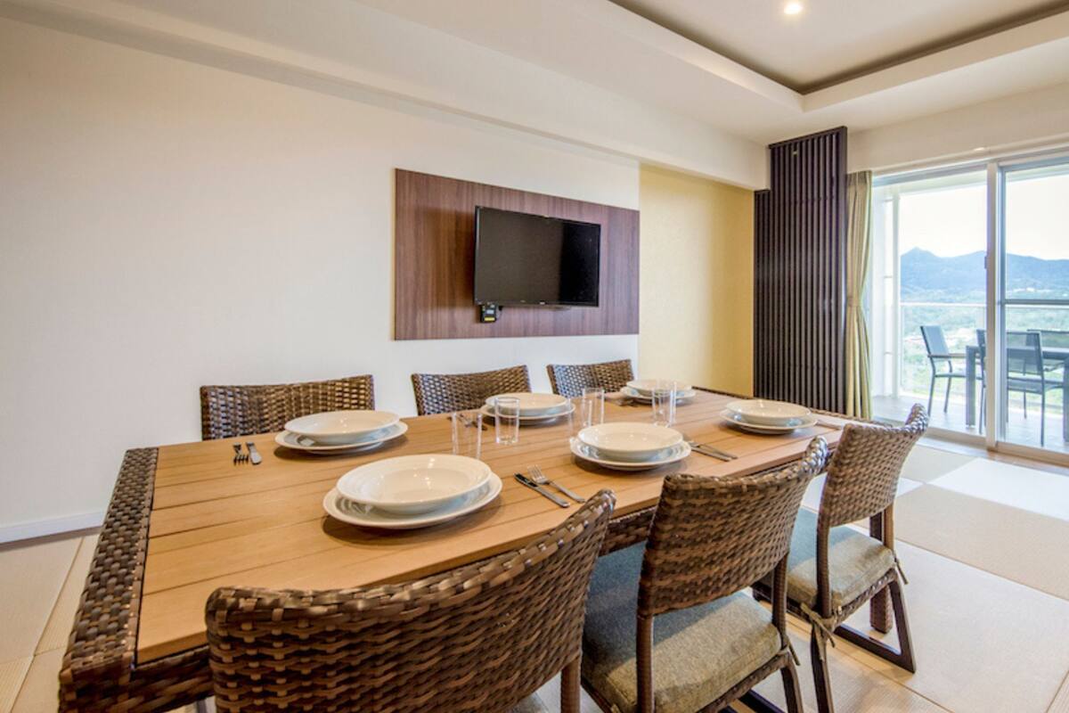 A dining area with a wooden table set for six, surrounded by wicker chairs, a flat-screen TV mounted on the wall, and a balcony with a view.