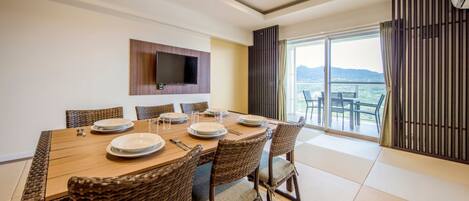 Un comedor con una mesa de madera dispuesta para seis personas, rodeado de sillas de mimbre, un televisor de pantalla plana en la pared y un balcón con vistas.