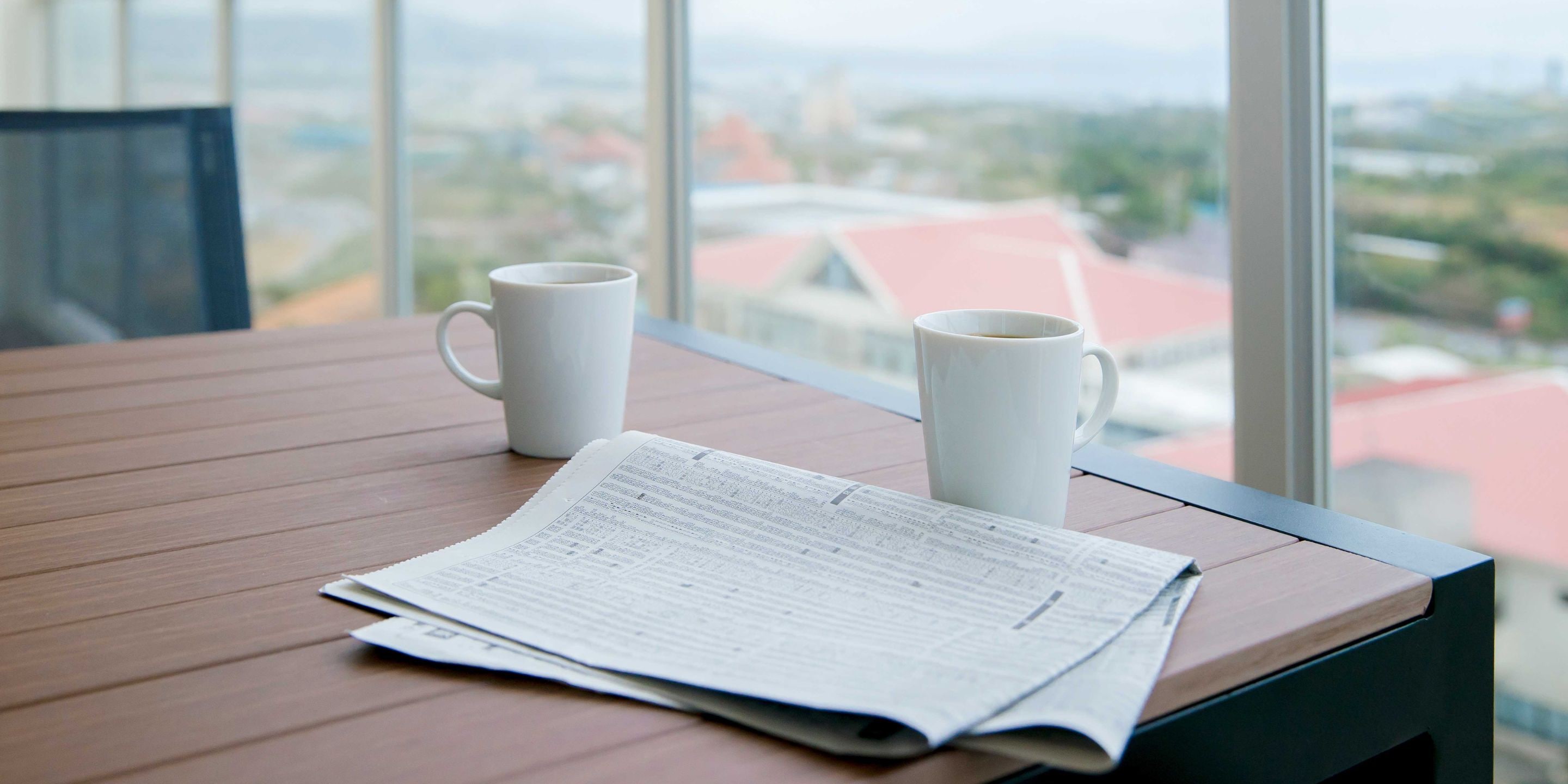 A newspaper on a wooden table with two white cups beside it, viewed through a window overlooking a cityscape.