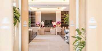 A modern hotel lobby with a central seating area, a flat-screen TV mounted on the wall, and potted plants.