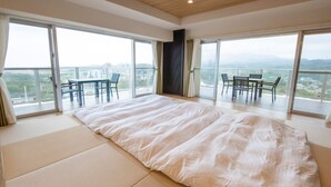 A spacious room with a large bed, a dining table, and chairs, and large sliding glass doors leading to a balcony with a city view. - Lieta Nakayama (Nago)