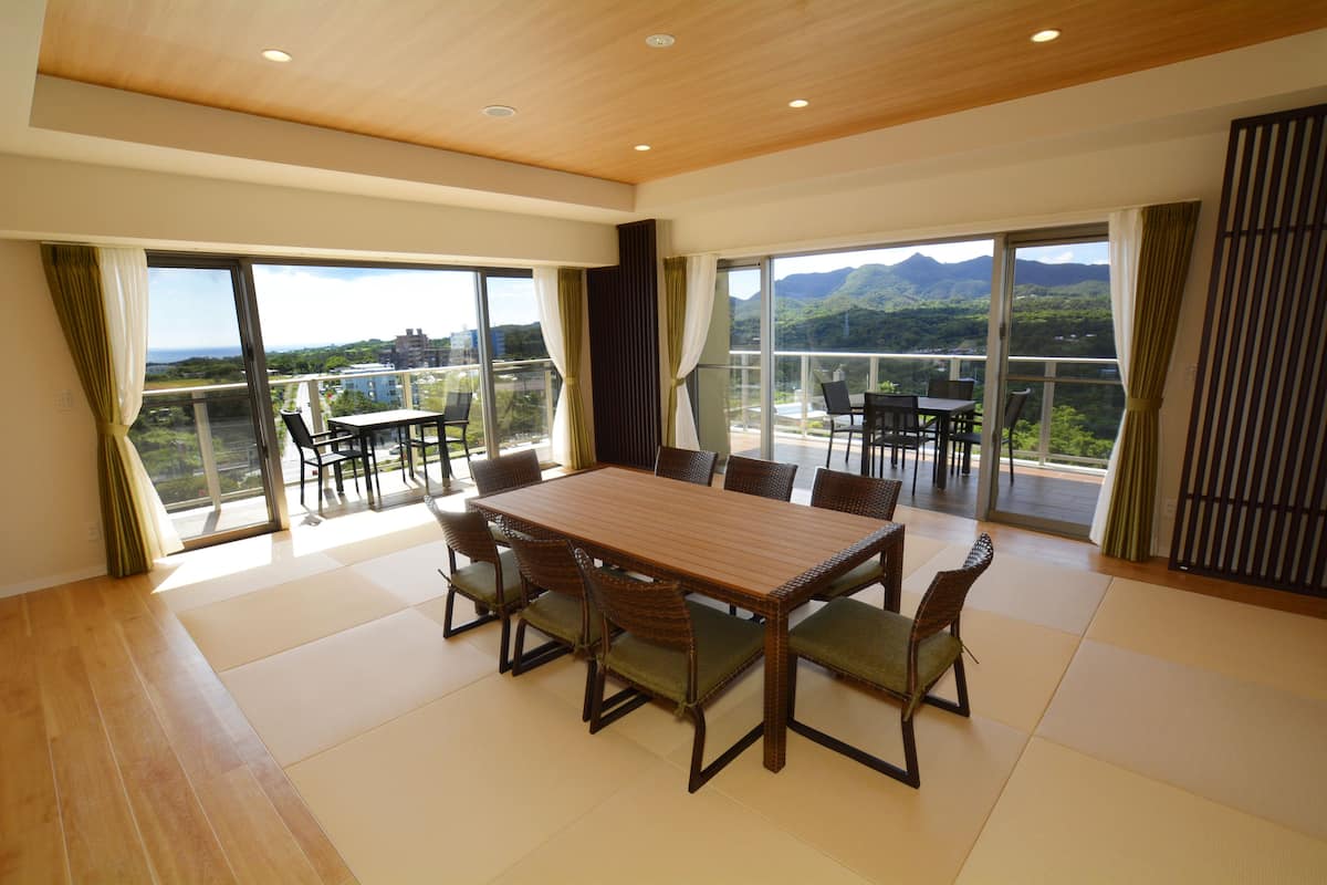 A spacious dining area with a large wooden table and chairs, large windows offering a view of mountains and greenery, and a balcony with outdoor seating.