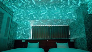 A room with a patterned wall, a bed with two pillows, a window with curtains, and a ceiling projection. - Lieta Nakayama (Nago)