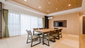 A modern dining area with a wooden table and chairs, a flat-screen TV mounted on the wall, and a large window with sheer curtains. - Lieta Nakayama (Nago)