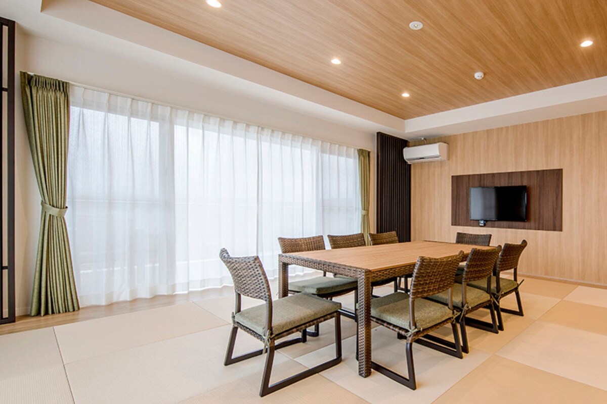 A modern dining area with a wooden table and chairs, a flat-screen TV mounted on the wall, and a large window with sheer curtains.