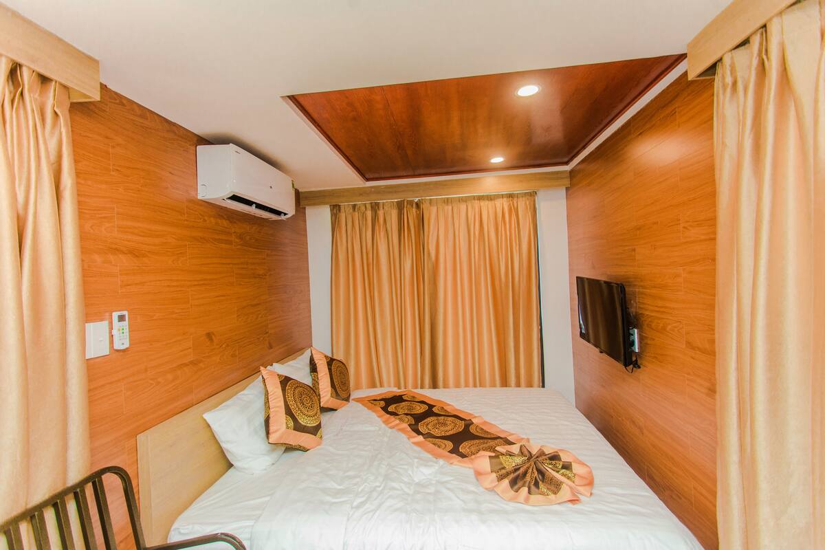 A hotel room with a wooden headboard, a bed with white bedding, a decorative throw, a wall-mounted TV, and an air conditioning unit.