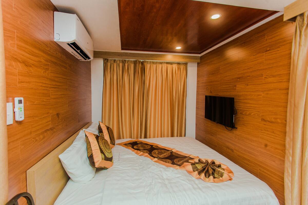 A hotel room with wooden walls, a bed with a patterned runner, a remote control, and a wall-mounted TV.