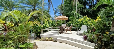 A tropical garden with a wooden deck, outdoor furniture, and a thatched umbrella.