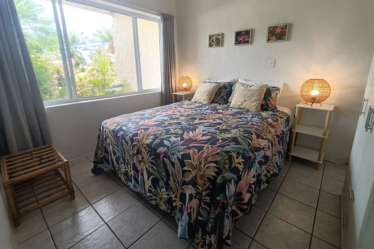 A bedroom with a large bed, two bedside lamps, a window with curtains, and a small wooden stool.