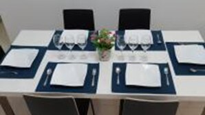 Une table à manger dressée pour quatre personnes, avec un vase de fleurs, des chaises noires et une nappe blanche.