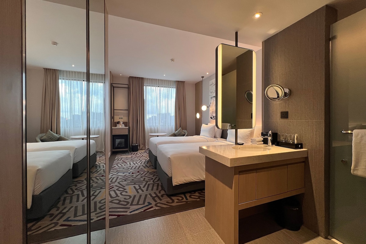 Ein Hotelzimmer mit zwei Betten, einem großen Spiegel, einem Waschbecken und einem gemusterten Teppich.