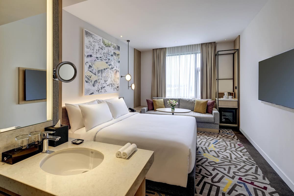 Habitación de hotel con una cama grande, una mesita de noche con espejo, una pequeña mesa redonda con un florero, un sofá, un televisor y un amplio ventanal con cortinas.