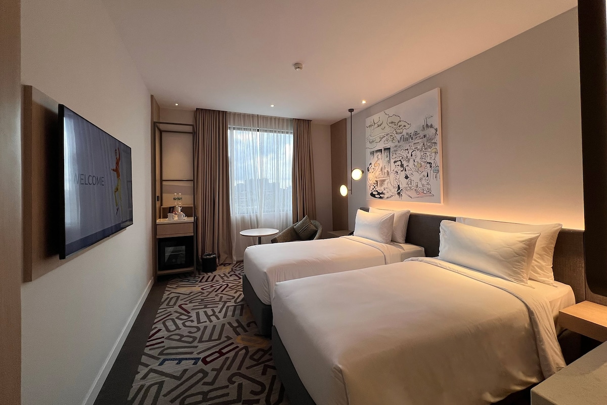 Ein Hotelzimmer mit zwei Betten, einem Fernseher, einem Schreibtisch, einem Stuhl, einem kleinen Tisch und einem Fenster mit Vorhängen.