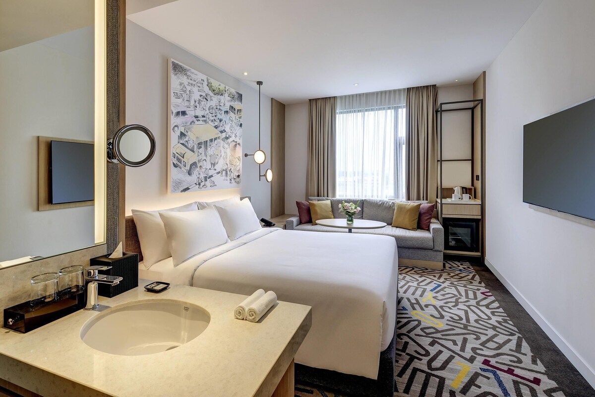 Ein Hotelzimmer mit einem großen Bett, einem Nachttisch mit Spiegel, einem kleinen runden Tisch mit Blumenvase, einer Couch, einem Fernseher und einem großen Fenster mit Vorhängen.