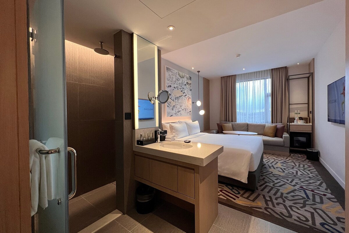 Ein Hotelzimmer mit einem großen Bett, einem Nachttisch, einem Spiegel, einem Badezimmer mit Dusche und einem gemusterten Teppich.