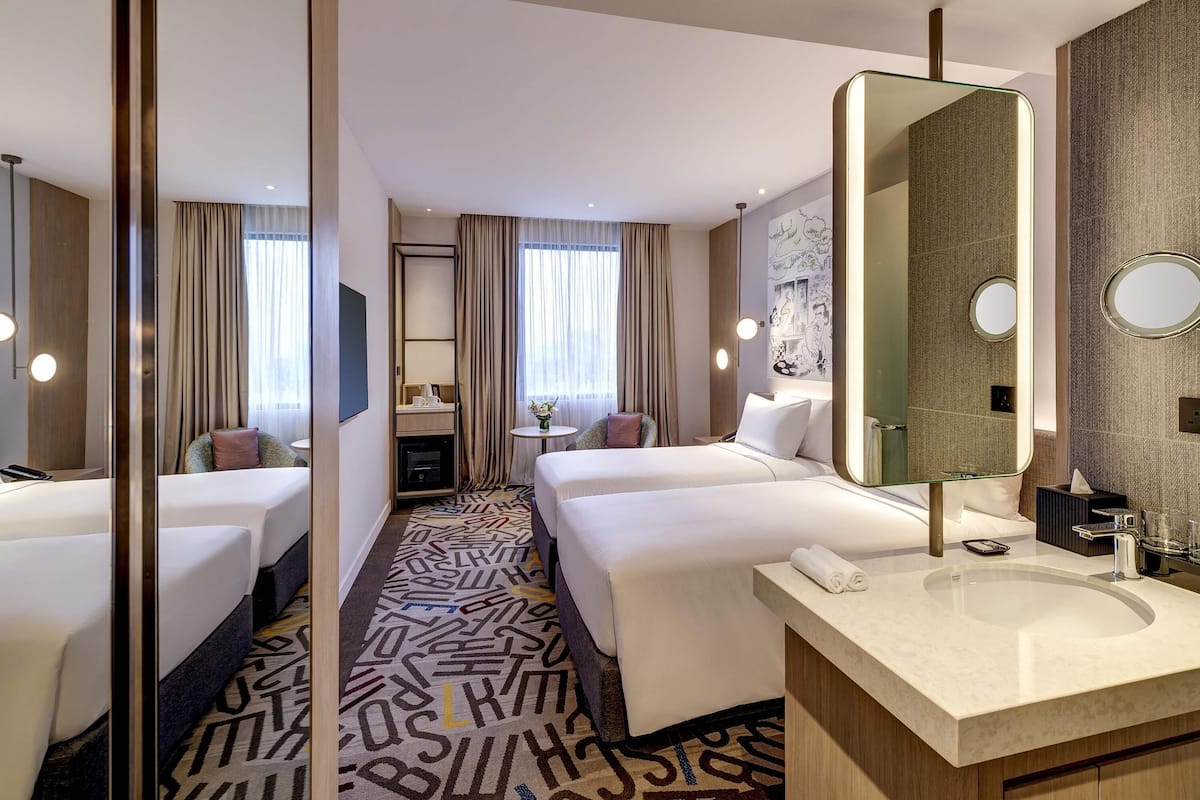 Habitación de hotel con dos camas, un espejo grande, un baño con lavamanos y una alfombra estampada.