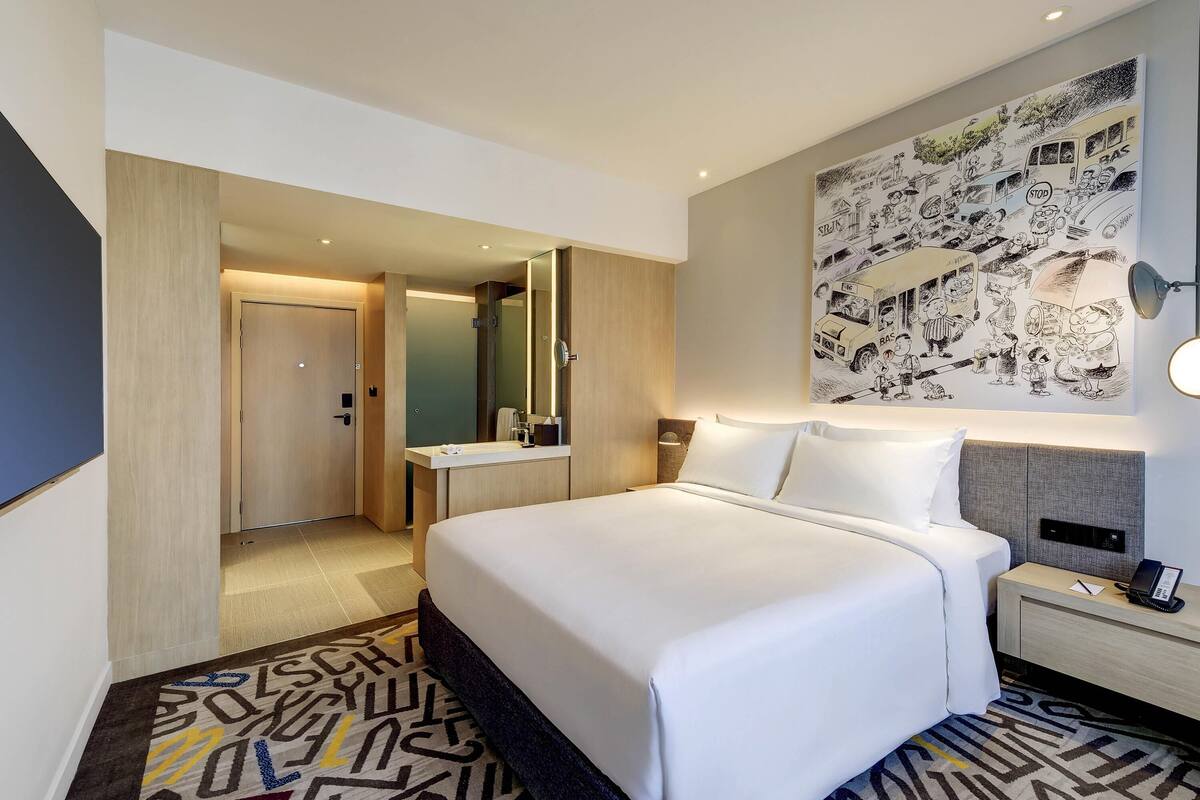 Habitación de hotel con una cama grande, una mesita de noche con teléfono, un espejo y una pared con un mural de caricaturas.
