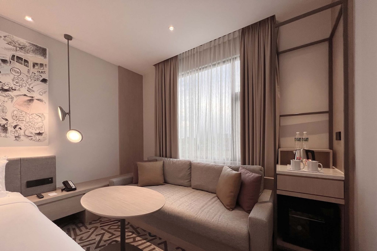 Ein modernes Hotelzimmer mit einer Couch, einem kleinen runden Tisch, einem Schreibtisch mit Lampe, einem Bett und einer Wand mit Wandmalerei im Comic-Stil.
