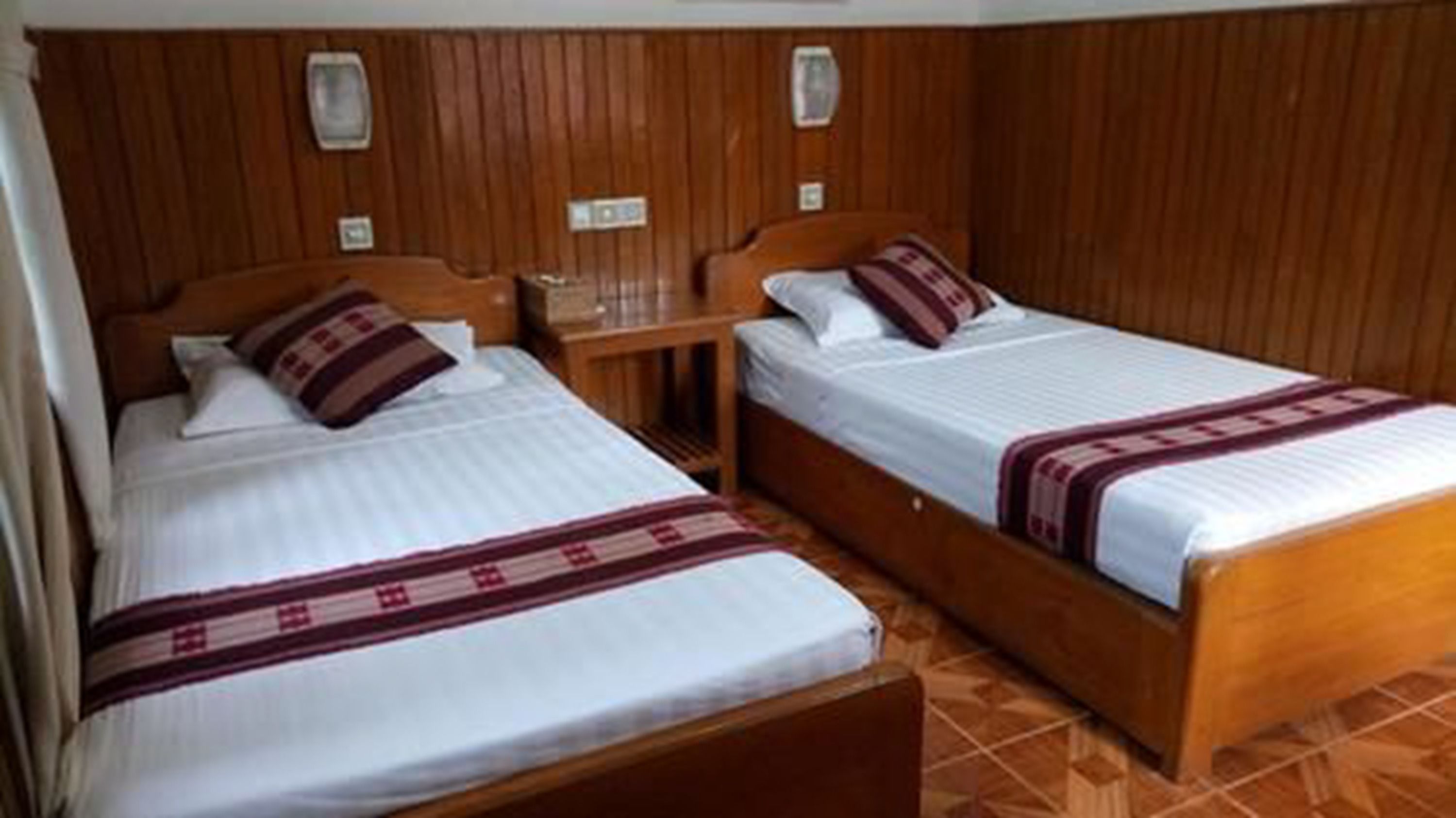 Dos camas individuales con cabeceros y mesitas de noche de madera, suelo de baldosas y paneles de madera en la pared.